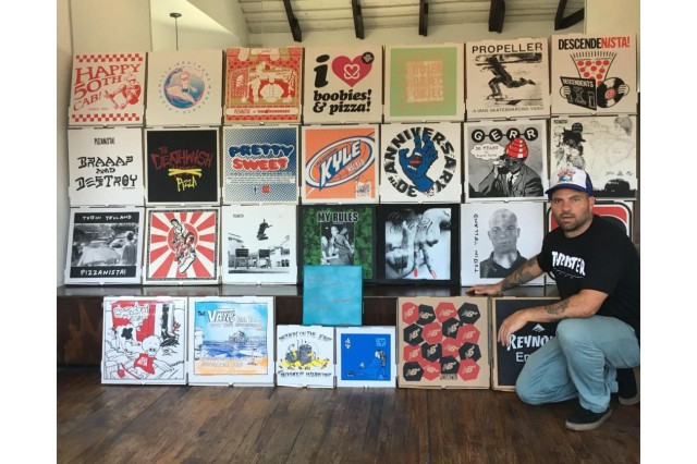 Man in baseball hat, black t-shirt, jeans & black sneakers, kneeling next to a wall of 25 decorative pizza boxes